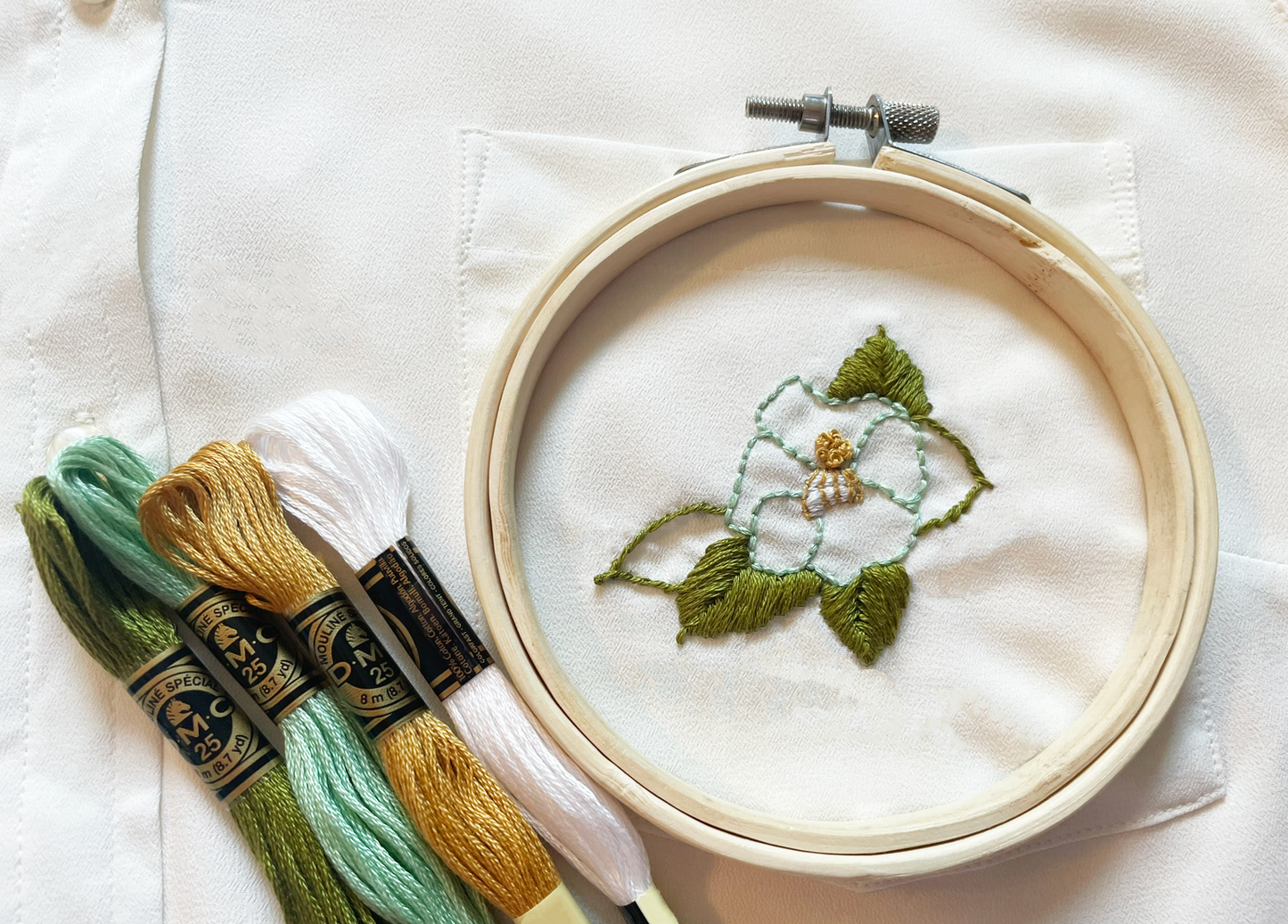 Closeup of a shirt pocket that has been embroidered with a turquoise camellia flower. A small embroidery hoop frames the completed design. Four skeins of embroidery floss, the colors used in the design, lie next to the hoop.