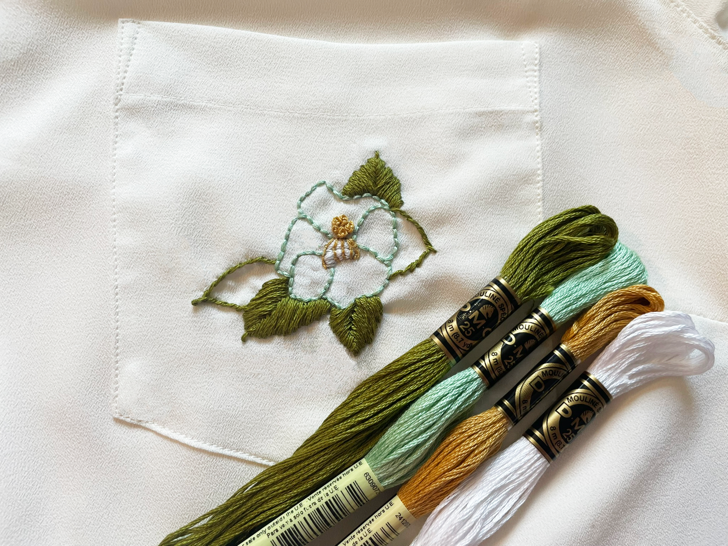 Closeup of an ivory shirt, the chest pocket has been embroidered with a turquoise camellia flower. The four embroidery floss skeins used to stitch the design are on the lower right corner.