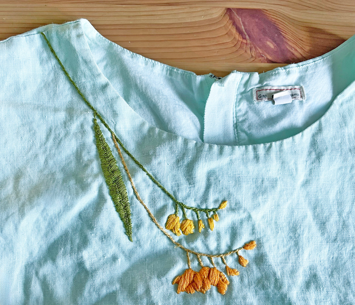 Closeup of turquoise dress hand embroidered along the neckline with yellow and orange blossoms, plus a large green leaf to the left side of the design.