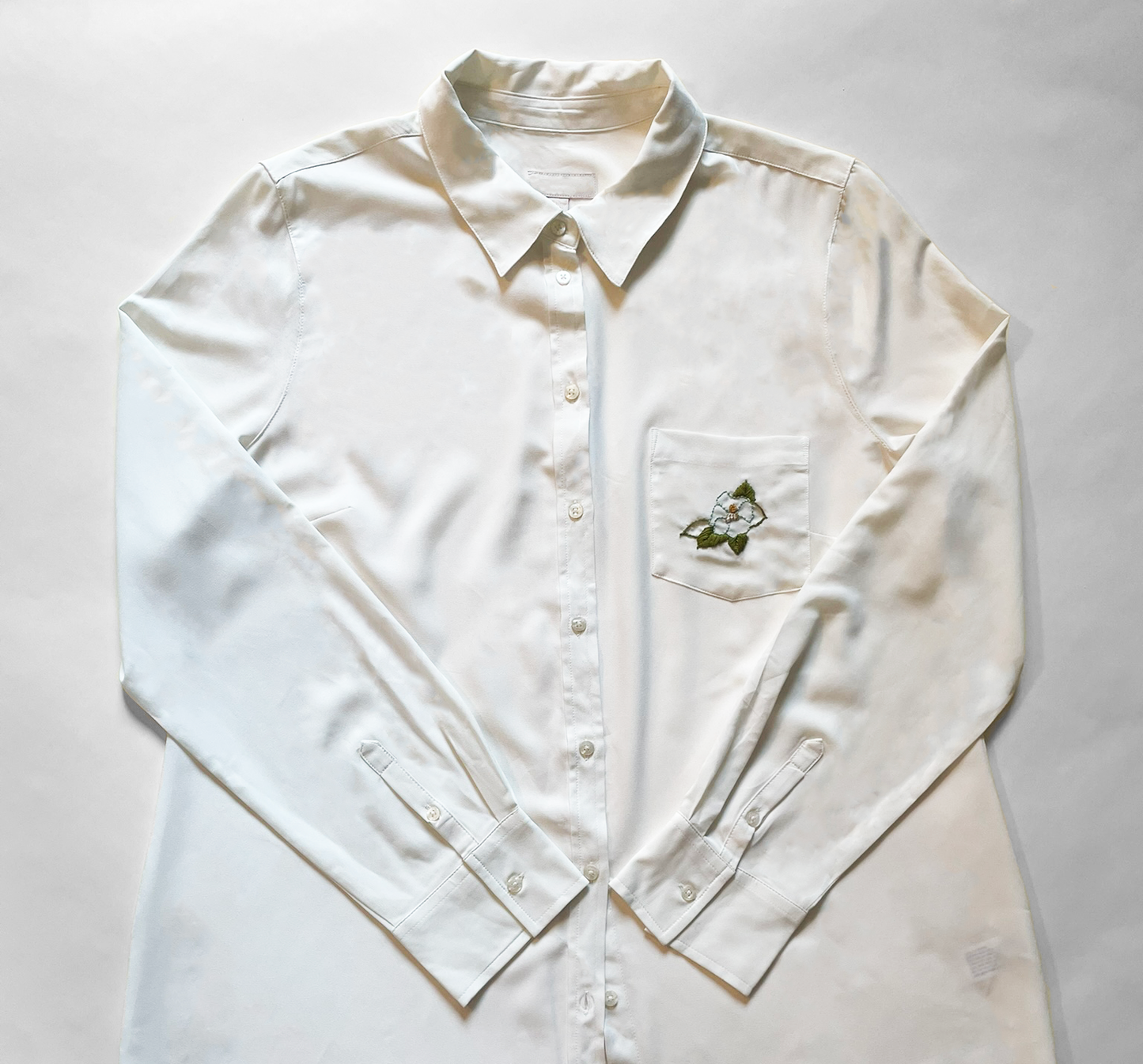 Hand embroidered ivory chiffon buttondown blouse lying on white ground. The chest pocket has been embroidered with a small turquoise camellia flower. Green leaves fan out underneath the petals.
