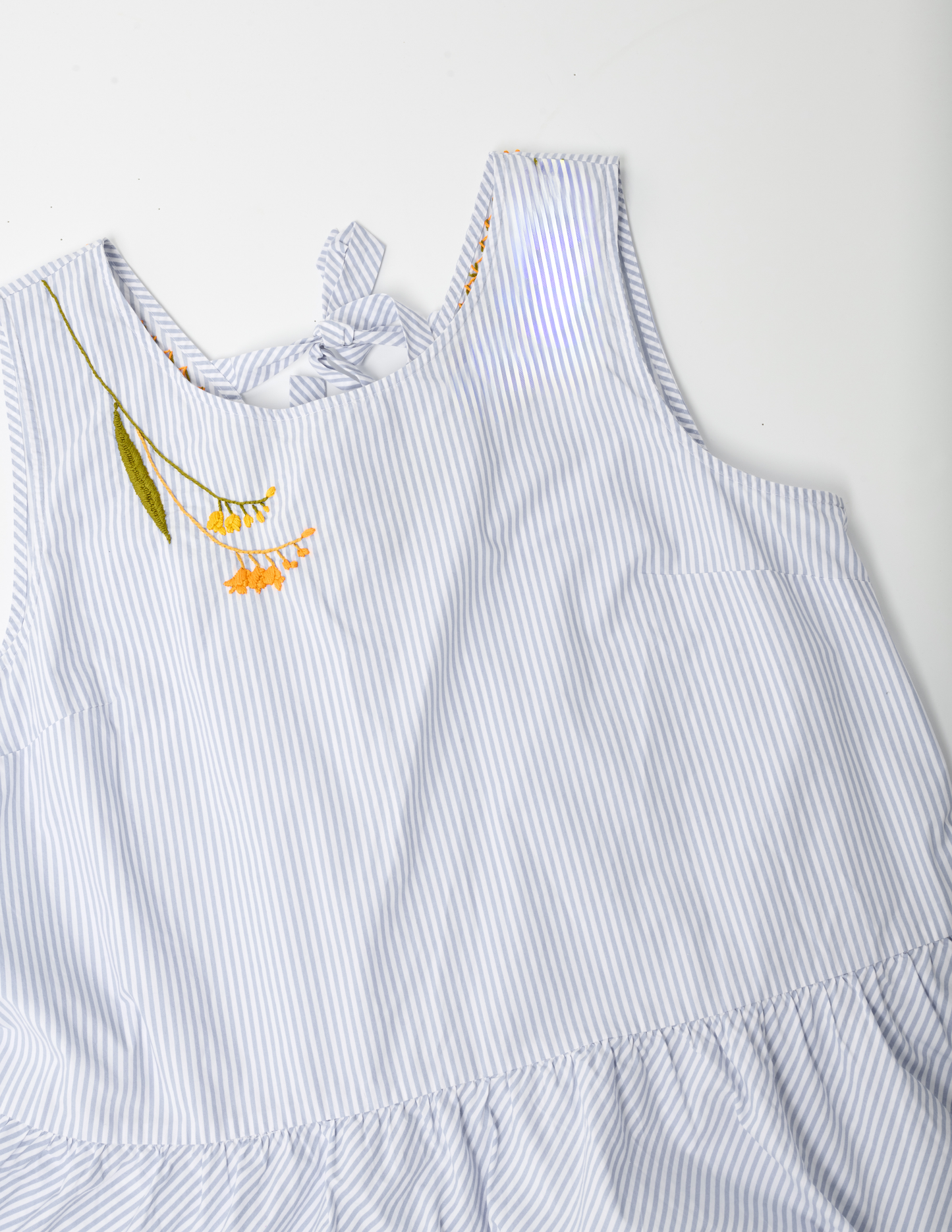 White and blue striped sleevelss peplum top on white ground. The right shoulder is hand embroidered with yellow and orange blossoms and a large olive green leaf on the left side of the design.