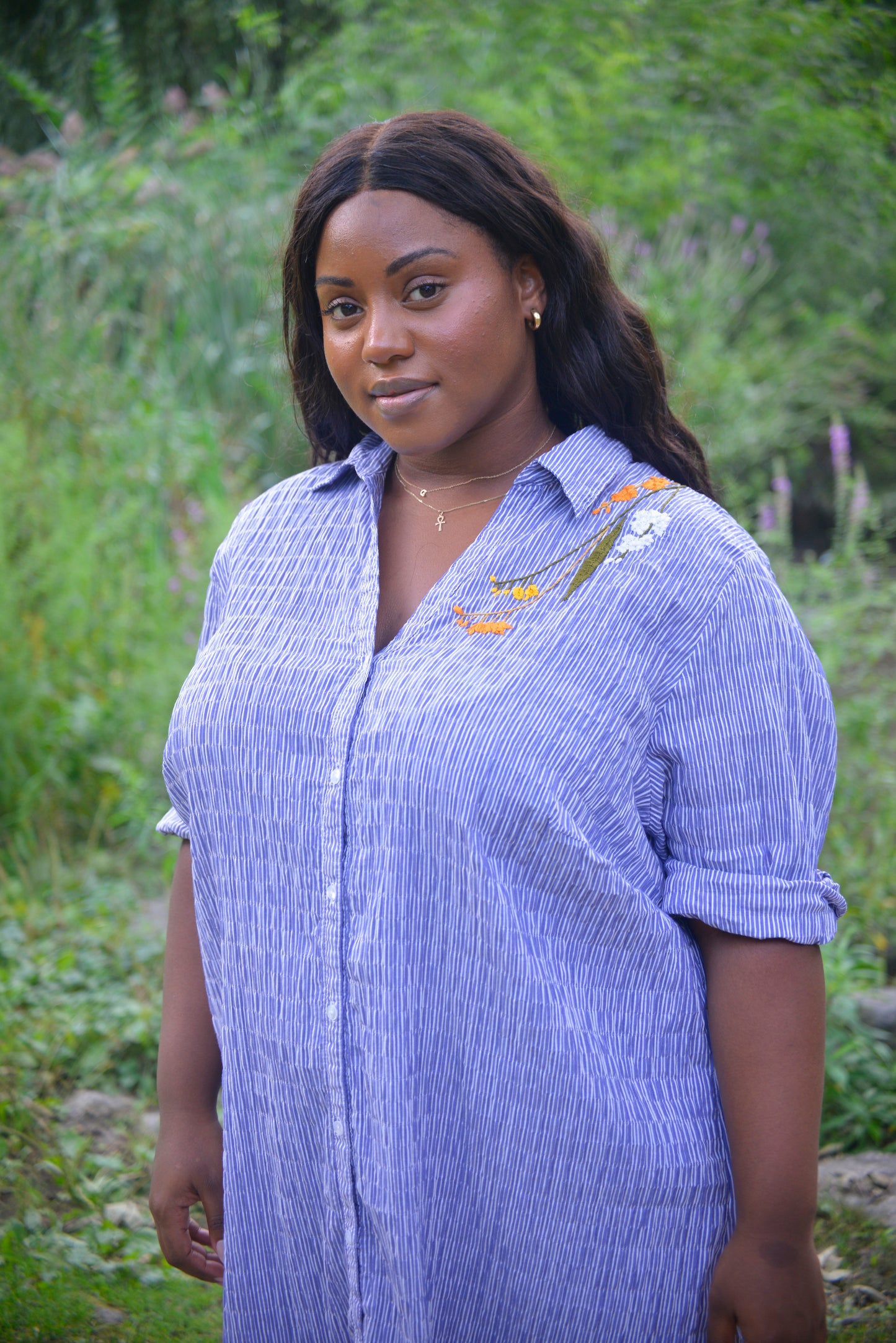 A curvy dark skinned model with long black wavy hair wears an oversized blue and white striped seersucker collared tunic. Pochoir.NYC hand embroidered orange, white, and gold blooms on the left shoulder.