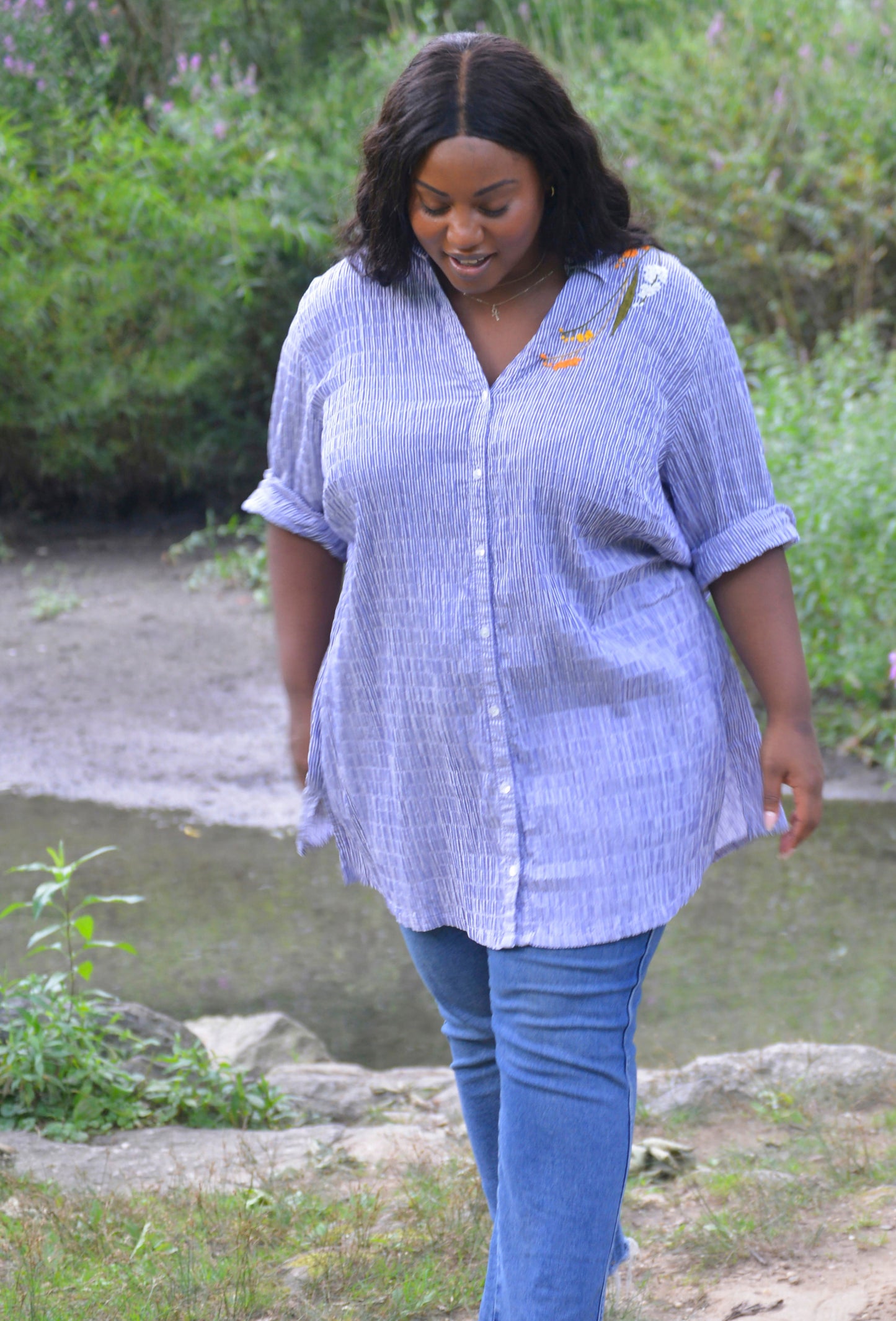 A curvy dark skinned model with long black wavy hair wears an oversized blue and white striped seersucker collared tunic. Pochoir.NYC hand embroidered orange, white, and gold blooms on the left shoulder.