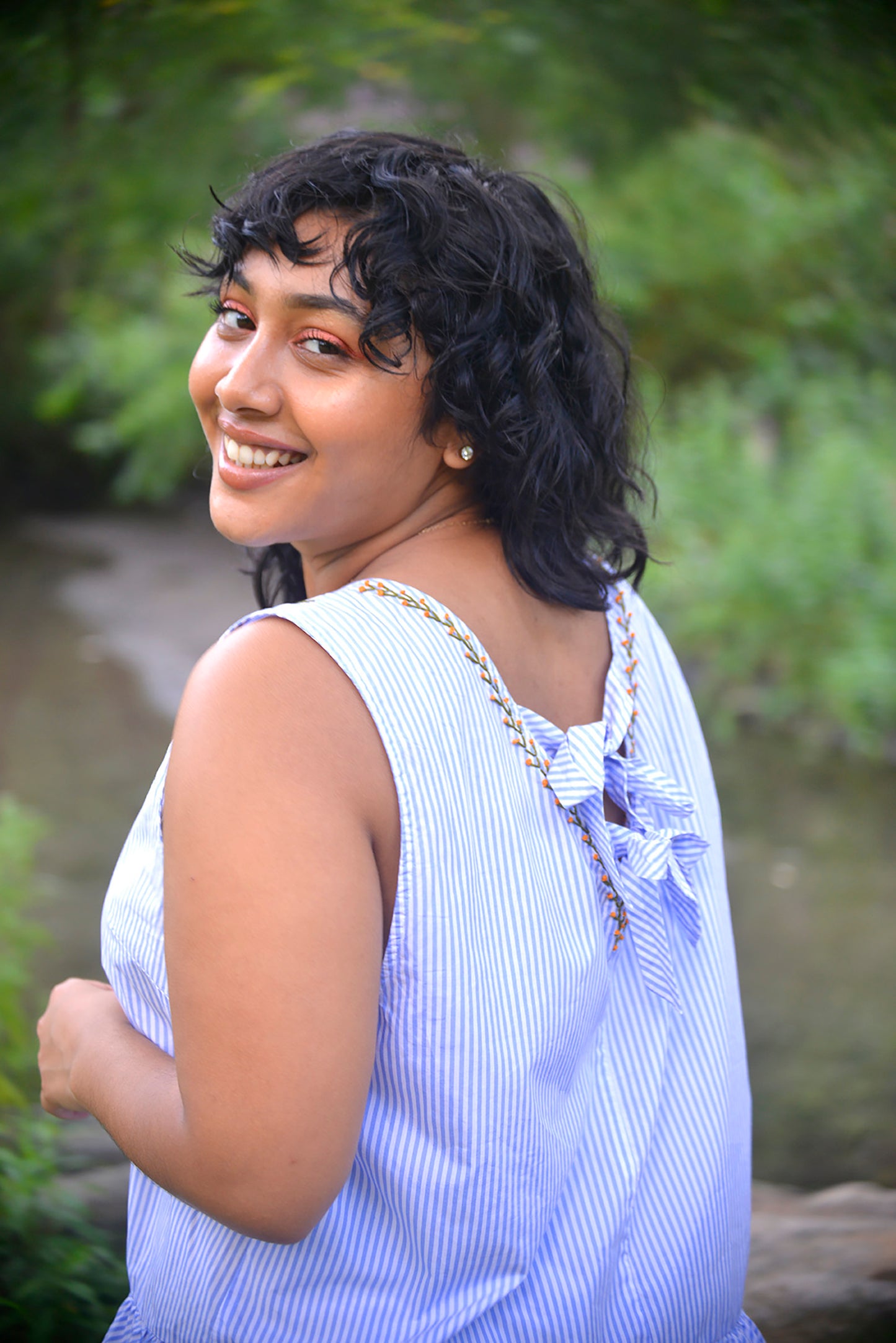 Model with dark hair and brown skin wears a blue and white striped peplum top. She peers over her shoulder to reveal that the back is closed with two ribbon ties and hand stitched with little orange blossoms down the neckline.
