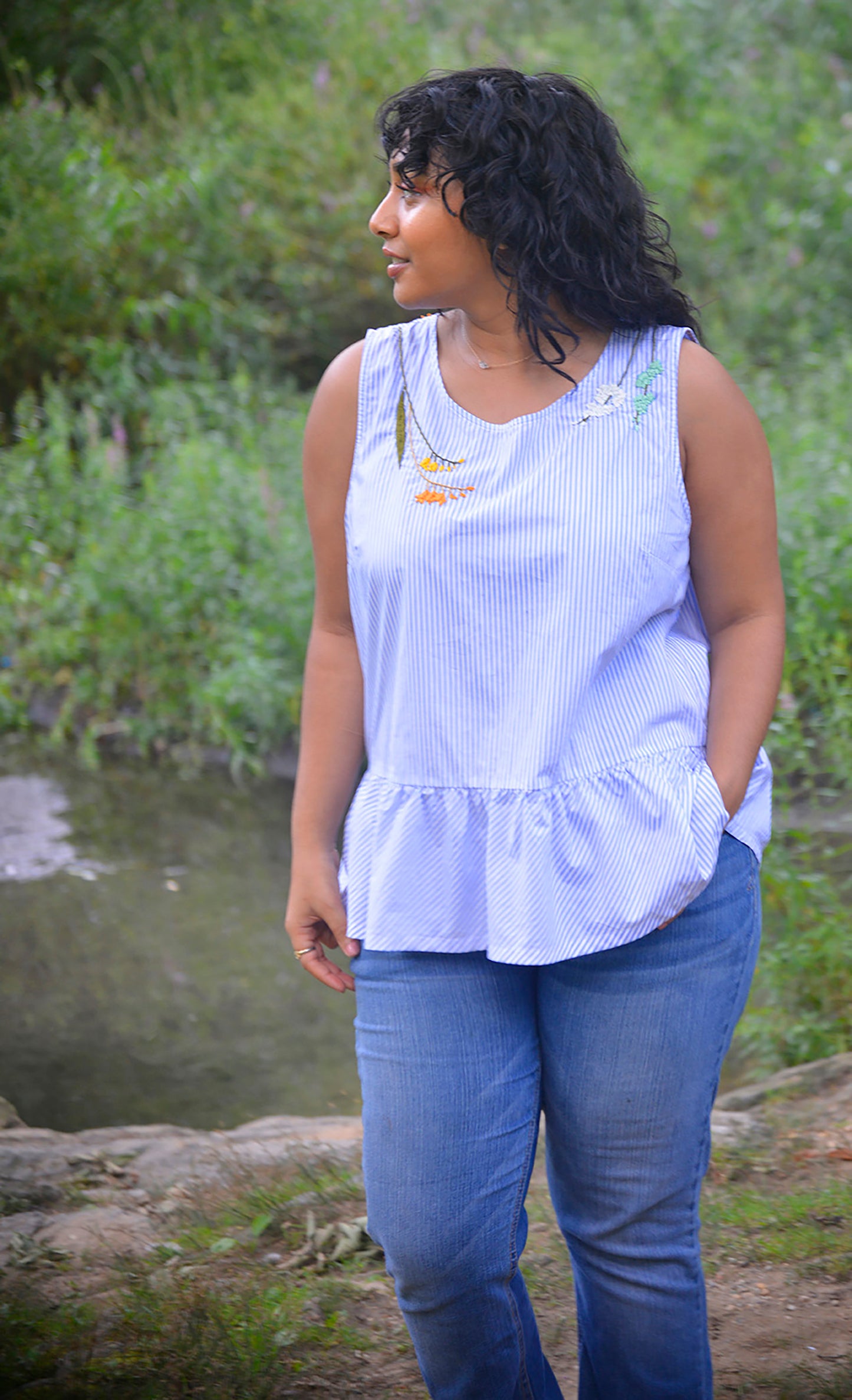 Model with dark hair and brown skin wears a blue and white striped peplum top hand embroidered by Pochoir.NYC with orange, yellow, white, and turquoise blossoms.