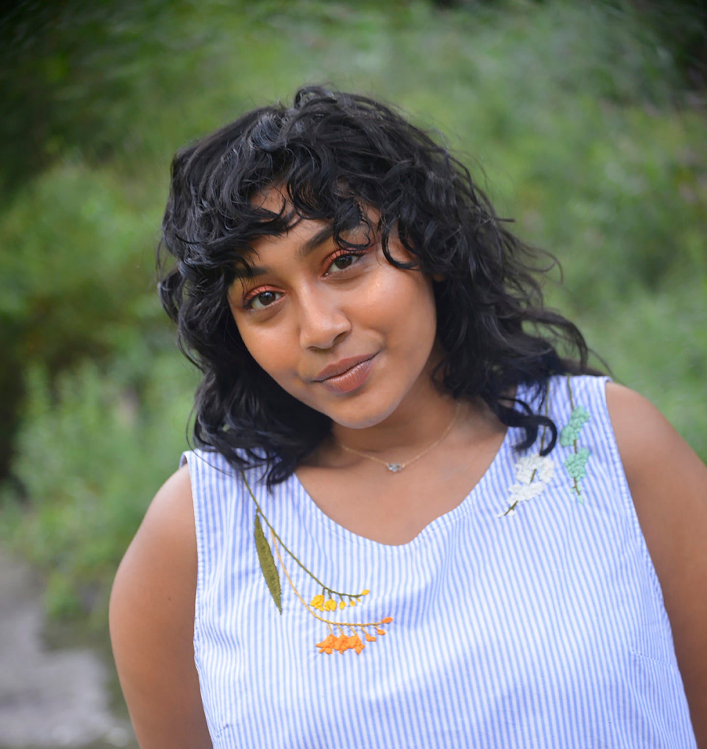 Model with dark hair and brown skin wears a blue and white striped peplum top hand embroidered by Pochoir.NYC with orange, yellow, white, and turquoise blossoms.
