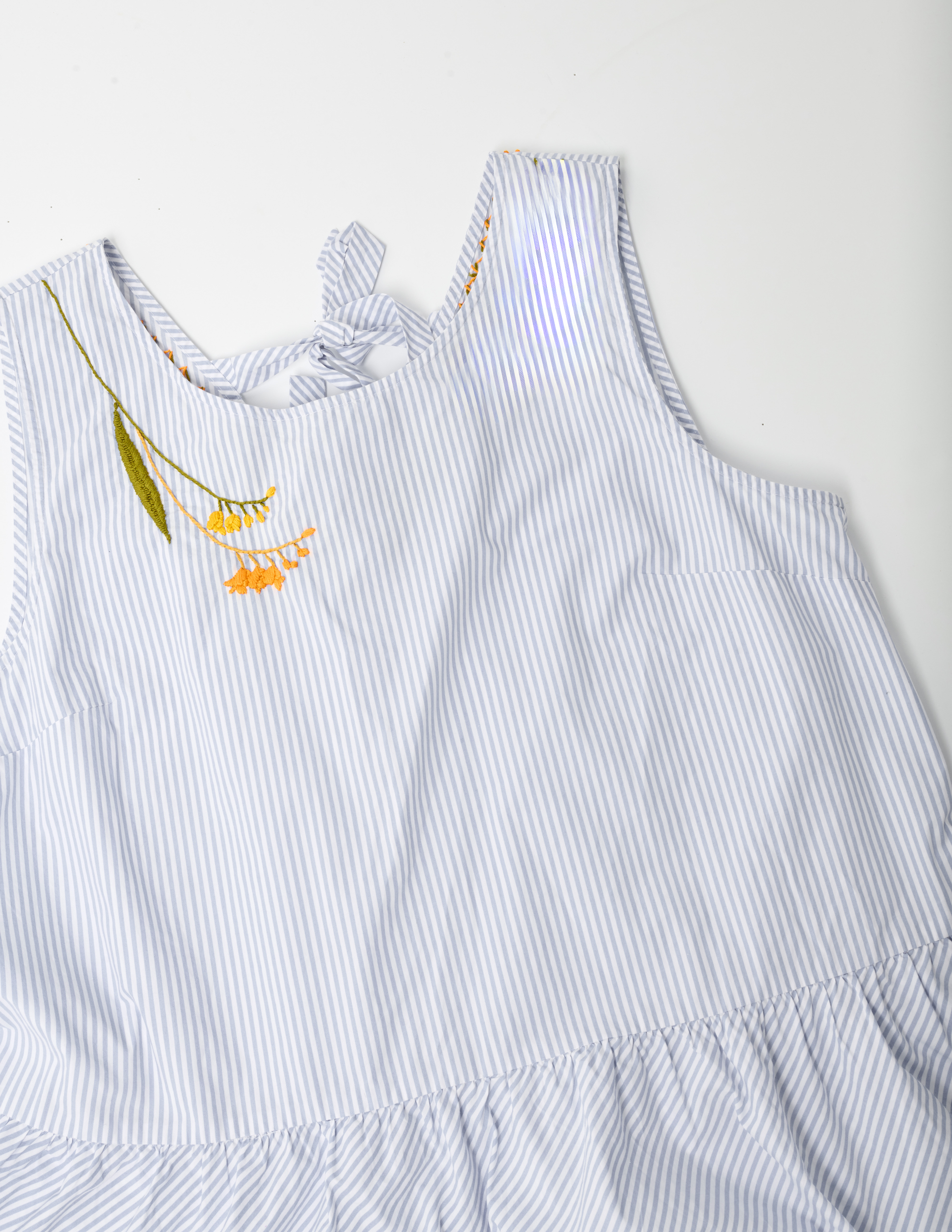 White and blue striped sleevelss peplum top on white ground. The right shoulder is hand embroidered with yellow and orange blossoms and a large olive green leaf on the left side of the design.