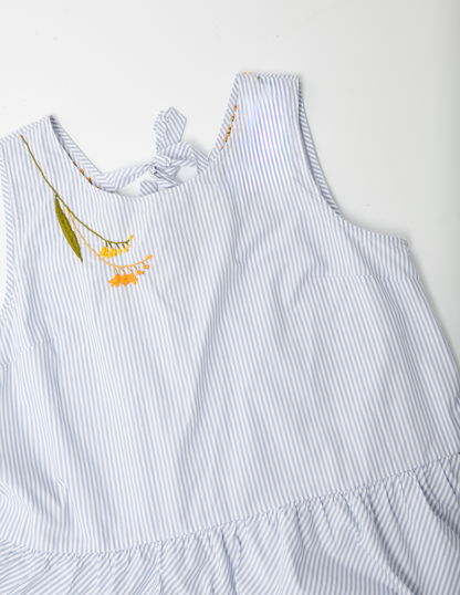 White and blue striped sleevelss peplum top on white ground. The right shoulder is hand embroidered with yellow and orange blossoms and a large olive green leaf on the left side of the design.
