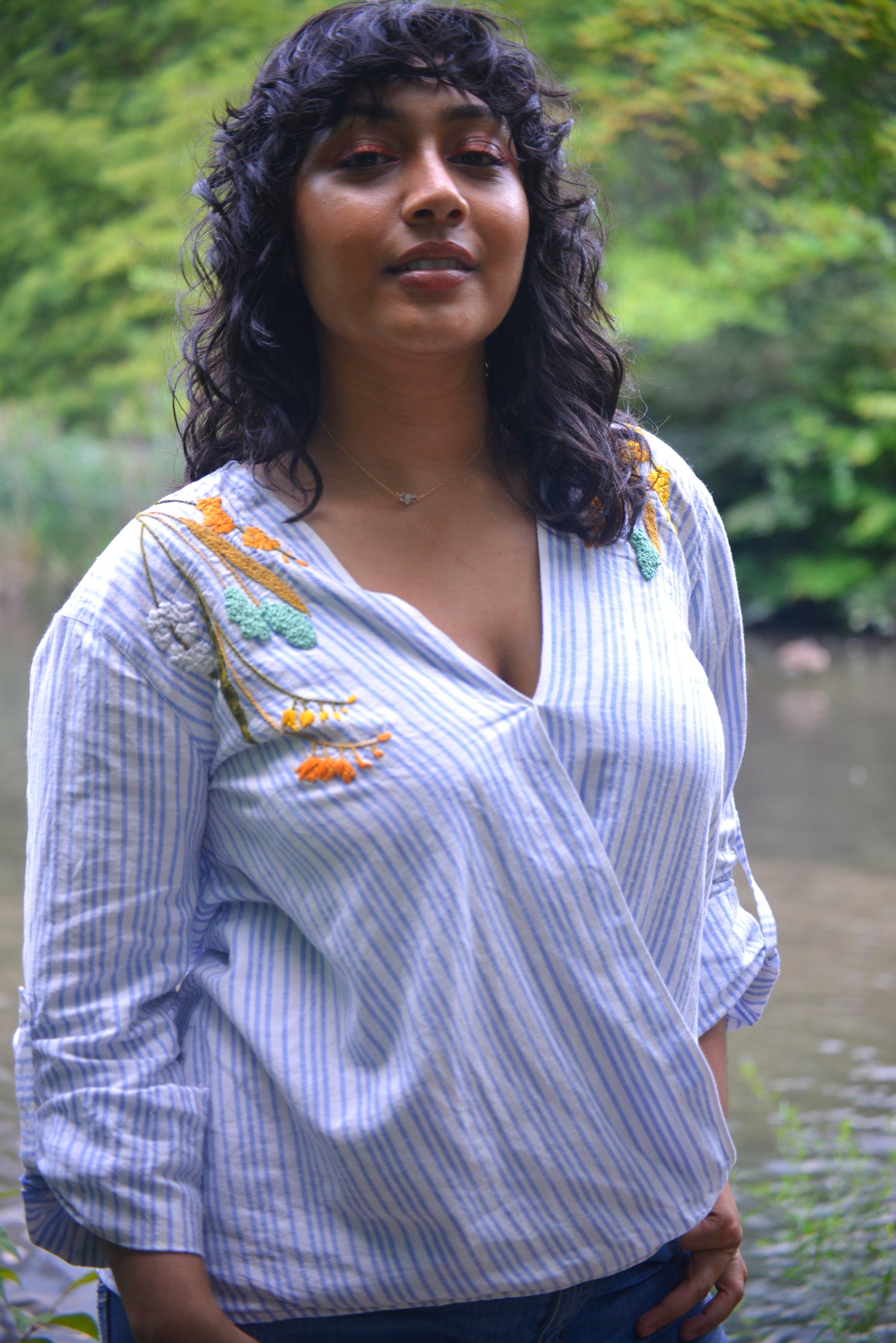 A thin model with brown skin and dark curly hair wears a blue and white striped linen wrap top hand embroidered by Pochoir.NYC with white, turquoise, yellow, and orange blossoms.