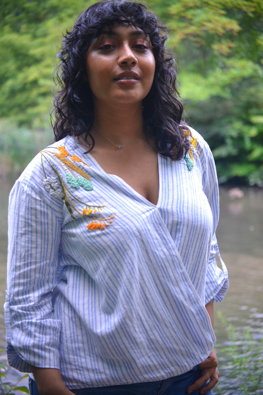 A thin model with brown skin and dark curly hair wears a blue and white striped linen wrap top hand embroidered by Pochoir.NYC with white, turquoise, yellow, and orange blossoms.