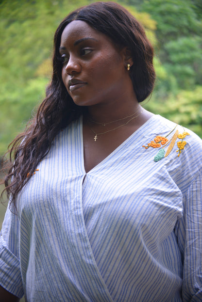 A curbymodel with dark brown skin and long dark curly hair wears a blue and white striped linen wrap top hand embroidered by Pochoir.NYC with white, turquoise, yellow, and orange blossoms.