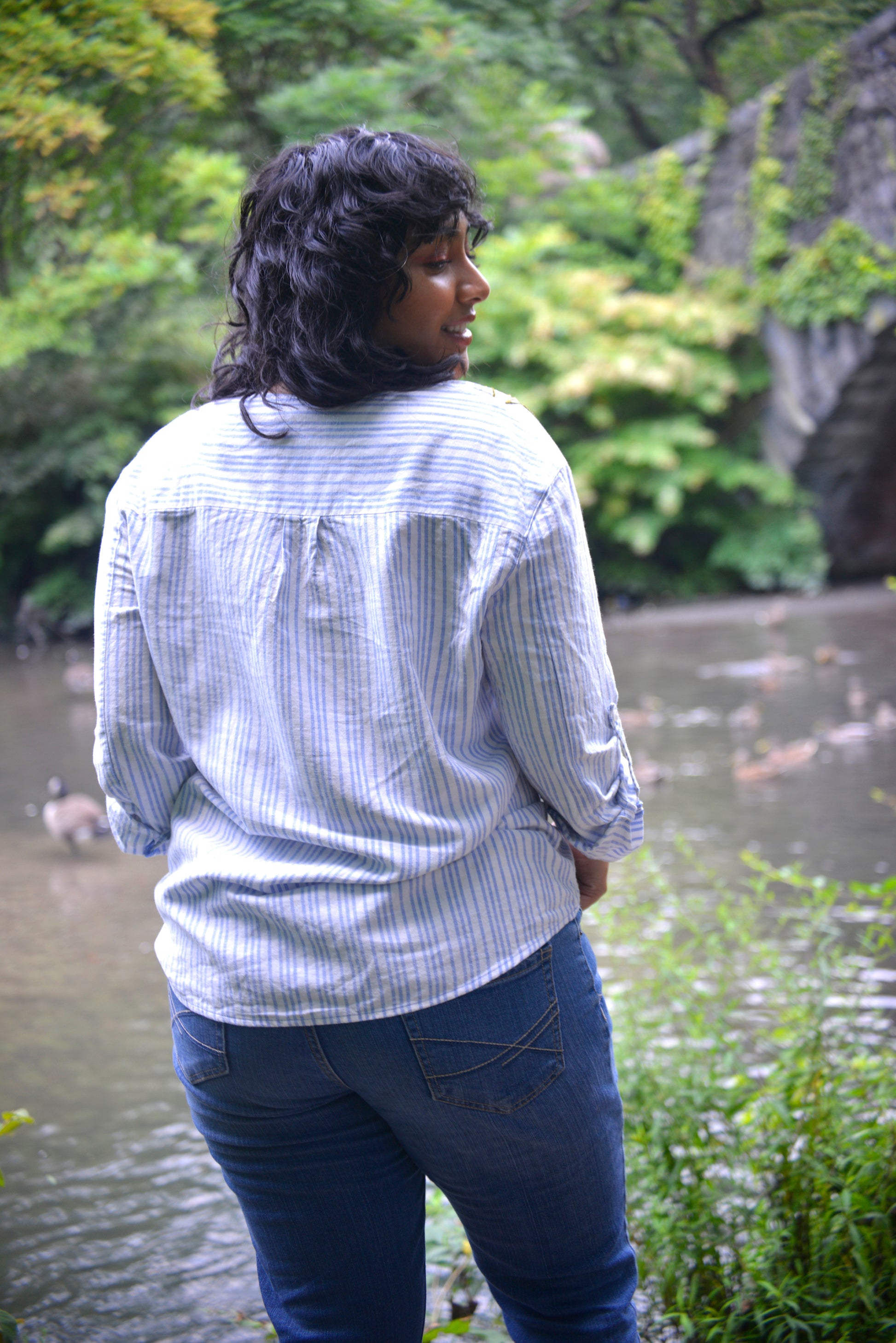 A thin model with brown skin and dark curly hair wears a blue and white striped linen wrap top. They are turned so their back is facing the camera and they're looking over their shoulder.