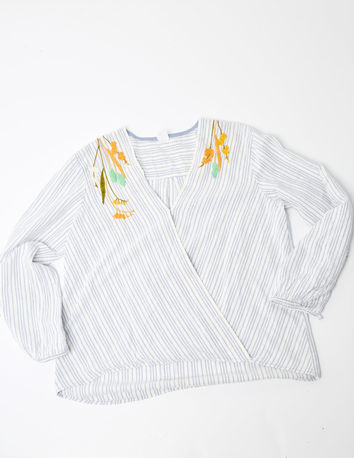 Image of a blue and white striped linen wrap top placed flat on a white background. Pochoir.NYC embroidered the neckline at the shoulders with orange, gold, turquoise, and white flowers.