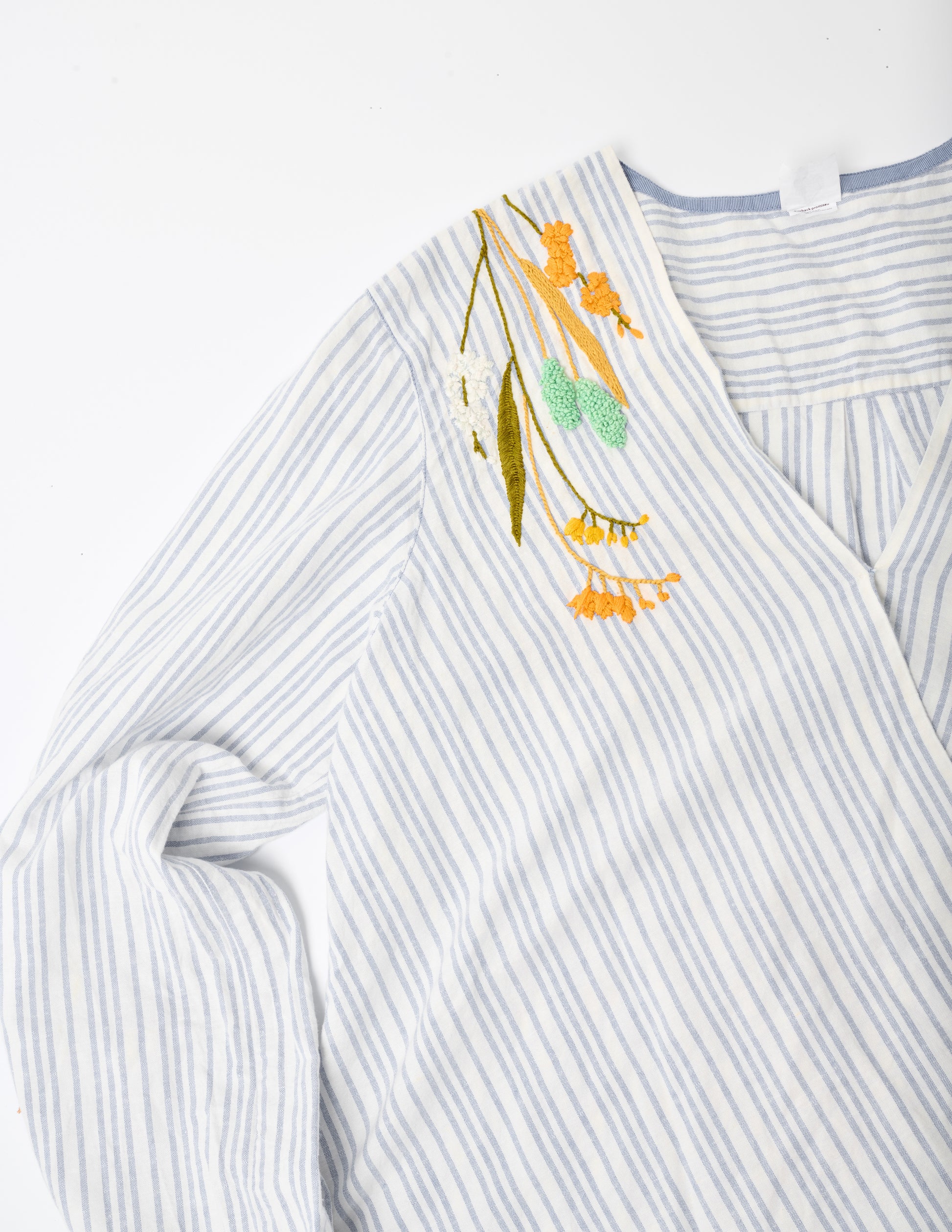 Closeup image of a blue and white striped linen wrap top placed flat on a white background. Pochoir.NYC embroidered the neckline at the shoulders with orange, gold, turquoise, and white flowers.