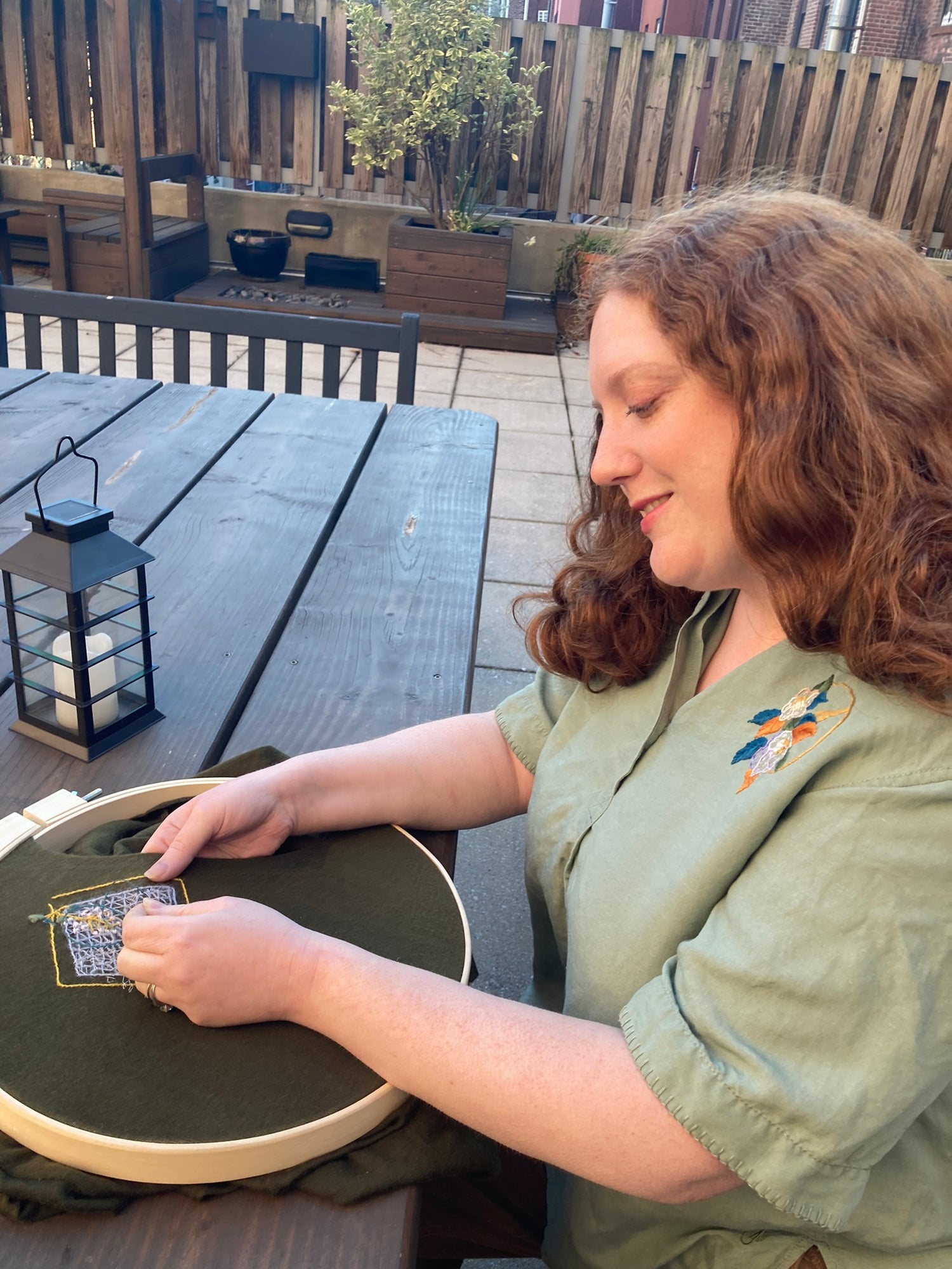 Photo of Ceci Zeichner, a red headed woman with pale skin, stitching a design onto a sweater while sitting on a patio. She is wearing a green linen tunic she hand embroidered with flowers.