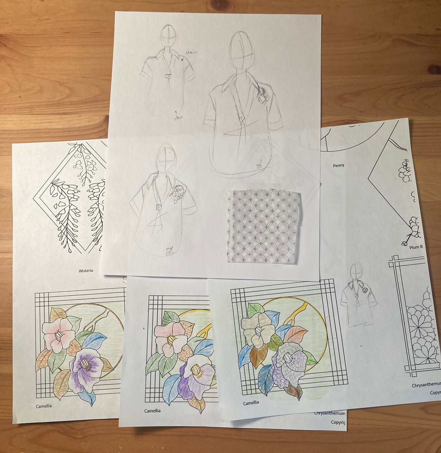 Image of several design sketches photographed against a wooden desk. Some sketches show a figure wearing an embroidered tunic. Others are colored in line drawings of flowers that will go on the shirt.