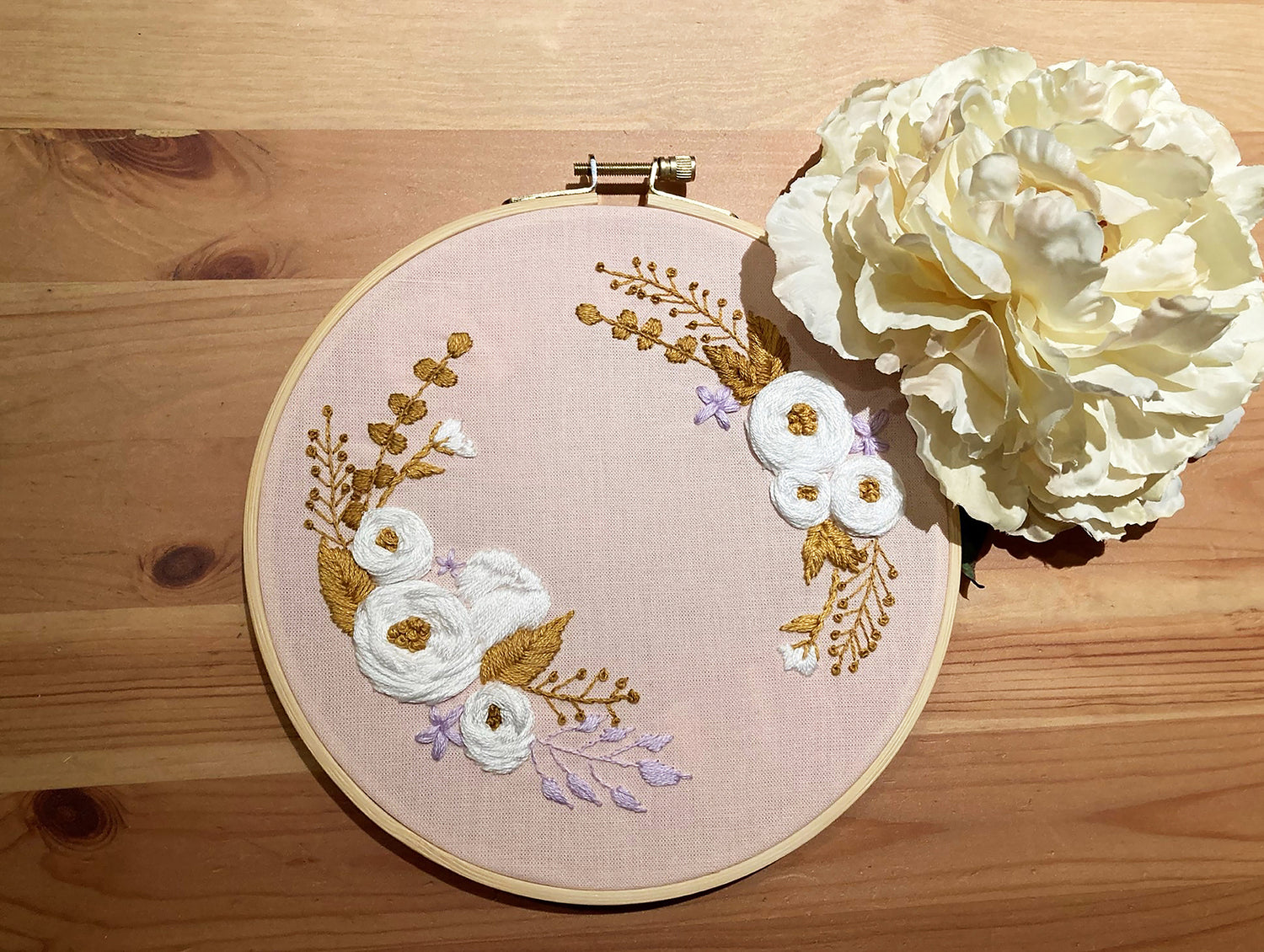 Photo of an embroidery hoop placed against a wooden desk. The Pochoir.NYC design includes white, gold, and lavender flowers stitched on pink cotton. There is a faux peony placed to the right of the hoop.