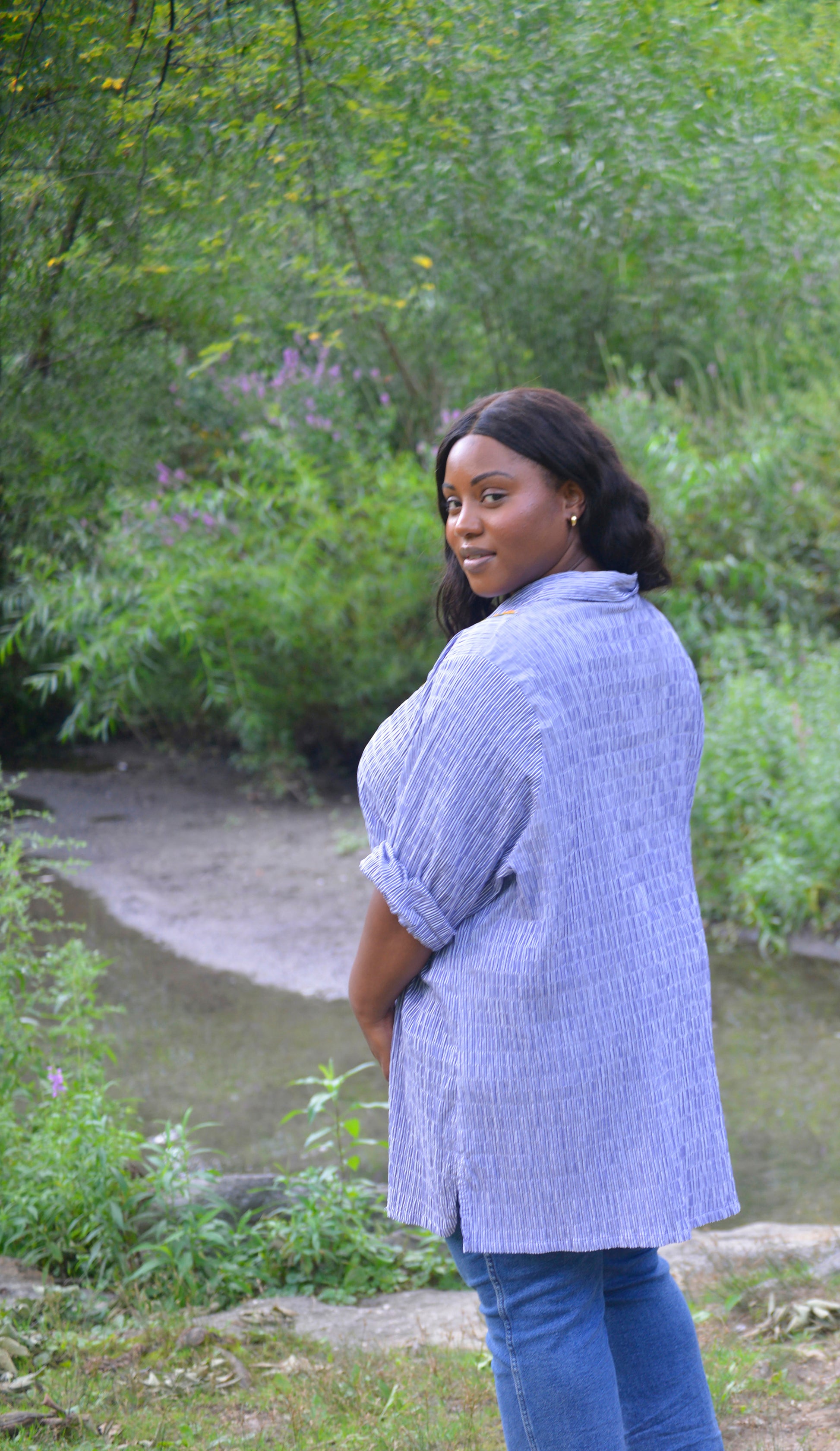 A curvy dark skinned model with long black wavy hair wears an oversized blue and white striped seersucker collared tunic. Pochoir.NYC hand embroidered the front with orange, white, and gold blooms on the left shoulder. The model stands with her back to the camera, looking over her shoulder, to show the viewer that the back is blank.