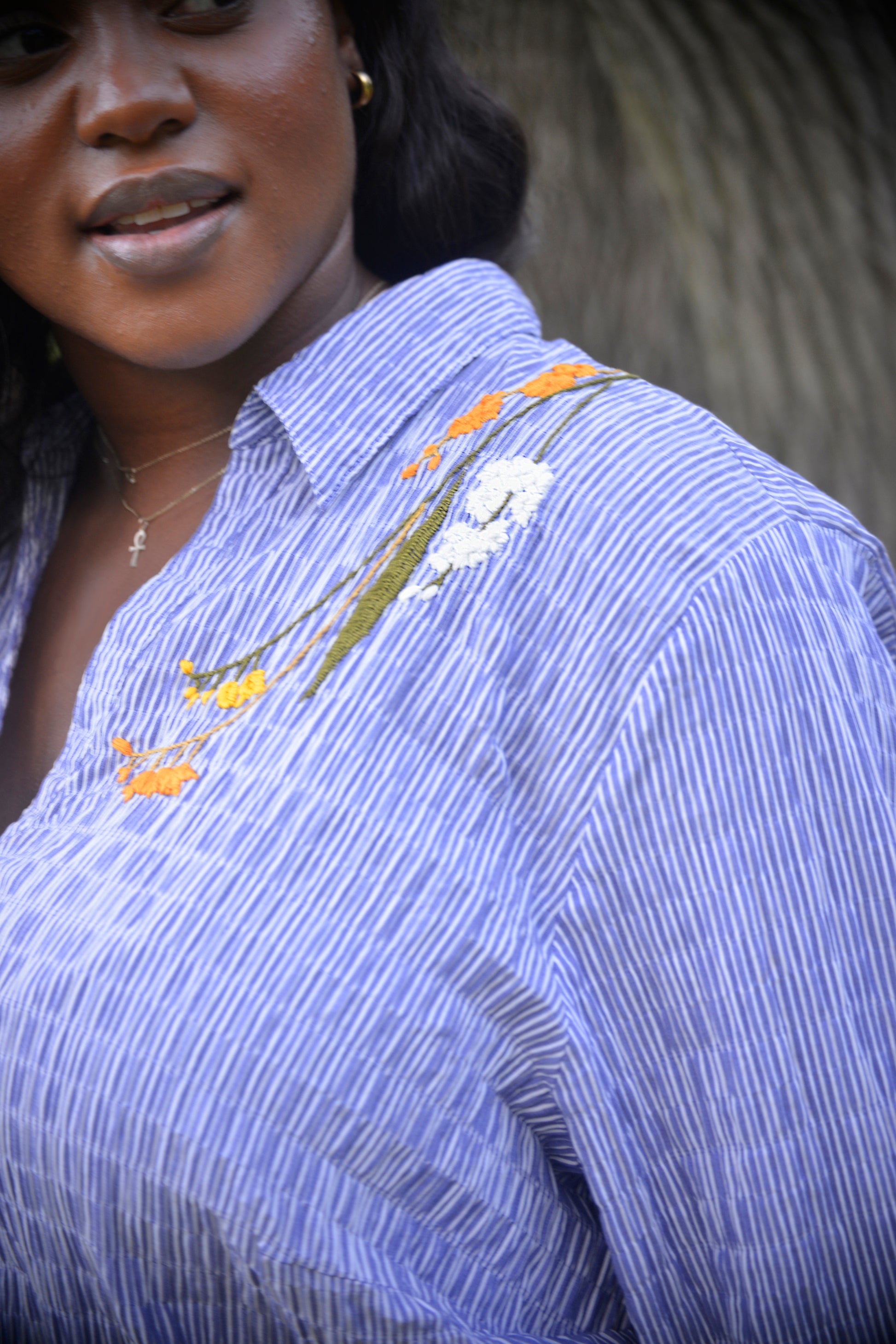 A curvy dark skinned model with long black wavy hair wears an oversized blue and white striped seersucker collared tunic. Pochoir.NYC hand embroidered orange, white, and gold blooms on the left shoulder.