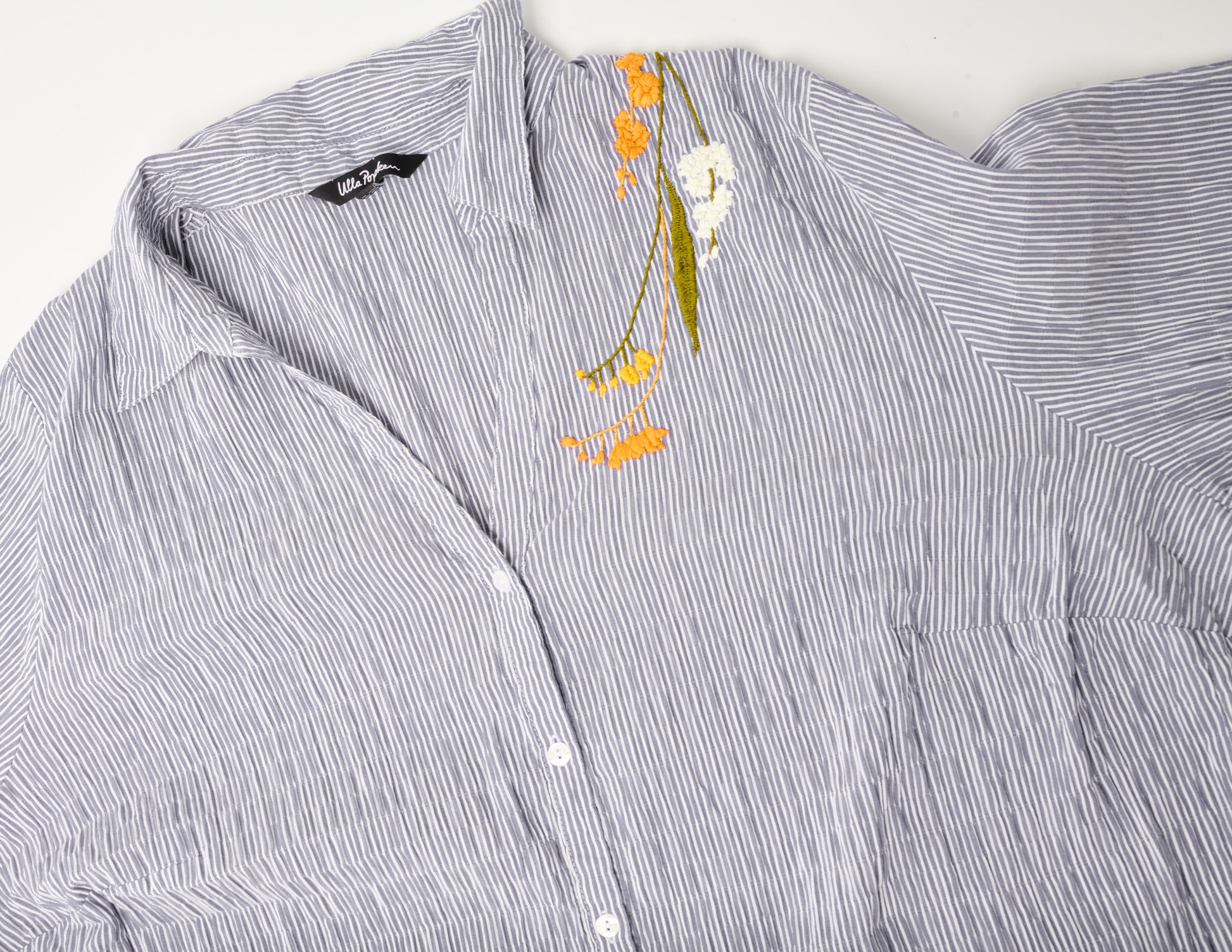 Closeup image of an oversized blue and white striped seersucker top. Pochoir.NYC embroidered the left shoulder with orange, white, and gold blossoms.