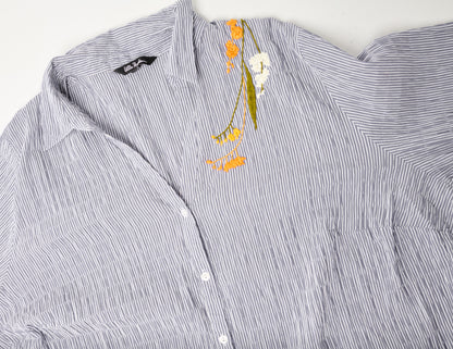 Closeup image of an oversized blue and white striped seersucker top. Pochoir.NYC embroidered the left shoulder with orange, white, and gold blossoms.