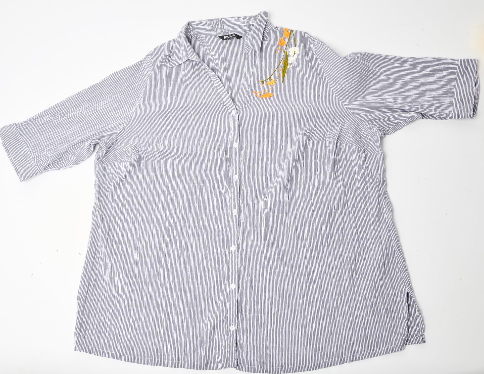 Flat image of an oversized blue and white striped seersucker top placed on a white background. Pochoir.NYC embroidered the left shoulder with orange, white, and gold blossoms.